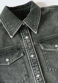 Ausgeblichene schwarze Denimjacke mit spitzem Kragen, Druckknöpfen in der Mitte und zwei aufgesetzten Brusttaschen, die mit Druckknöpfen gesichert sind.