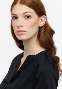 Blusa nera con scollo a V, realizzata in tessuto morbido e con una struttura sottile. Il modello ha capelli rossi ondulati e un'espressione neutra.