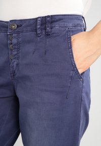 Close-up van de hand van een persoon in de voorzak van een blauwe broek met knopen, gedragen met een witte top.