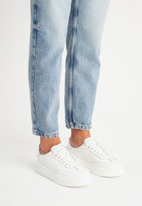 Białe sneakersy z teksturowaną skórzaną cholewką, płaskimi sznurowadłami i masywną gumową podeszwą. Noszone z jasnoniebieskimi, lekko przetartymi dżinsami.