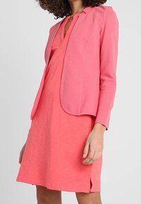 Kobieta w jasnoróżowej sukience do kolan z dekoltem w serek i dopasowanym żakiecie, stojąca na tle jednolitego jasnego tła.