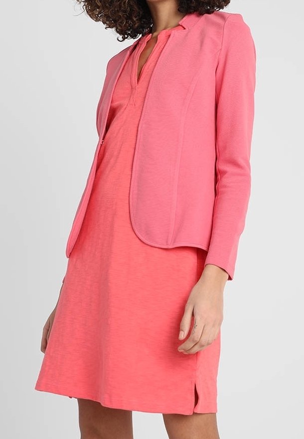 Kobieta w jasnoróżowej sukience do kolan z dekoltem w serek i dopasowanym żakiecie, stojąca na tle jednolitego jasnego tła.