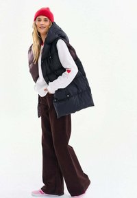 Žena v červenej čiapke, bielej mikine s detailom srdca, čiernej prešívanej veste, hnedých širokých nohaviciach a ružových teniskách stojaca proti bielemu pozadiu.