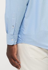 Camicia azzurro chiaro a maniche lunghe con polsini abbottonati, caratterizzata da un tessuto liscio e una vestibilità sartoriale. Cuciture visibili e dettaglio dell'orlo curvo.