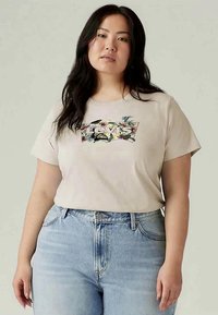 Mulher com cabelo longo e escuro, vestindo uma t-shirt bege da Levi's com um logótipo floral e jeans de cintura alta azul claro, de pé contra um fundo simples.