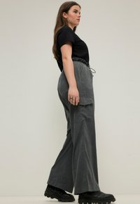 Housut, joissa on leveät lahkeet ja jotka on valmistettu teksturoidusta kankaasta. Niissä on kiristettävä vyötärö. Yhdistetty mustan lyhythihaisen t-paidan ja paksujen mustien kenkien kanssa.