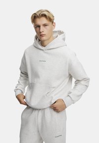 Grå hoodie lavet af en bomuldsblanding, med en frontlomme og ribbede manchetter. Mærket "Caspara" er trykt i grønt på brystet.