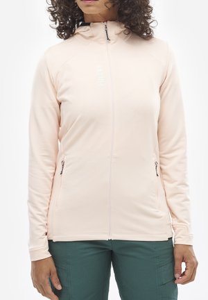 Femme portant une veste zippée rose clair à capuche et un pantalon vert, debout les mains reposant le long du corps.