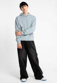 Jasnoniebieska bluza z kapturem z przednią kieszenią i nadrukowanym wzorem, połączona z czarnymi spodniami o szerokim kroju z bocznym łańcuszkiem oraz białymi butami sportowymi.