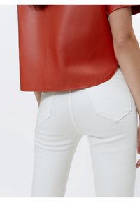 Top corto de cuero rojo de manga corta combinado con jeans blancos de corte ajustado que tienen bolsillos traseros y una textura suave. El atuendo presenta un contraste en color y material.