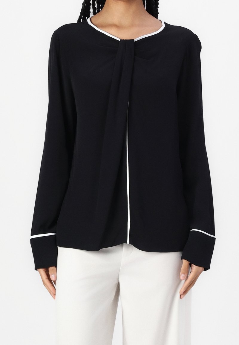 Blouse noire à manches longues avec un nœud au niveau du décolleté, bord blanc le long du col et des poignets, texture lisse et coupe décontractée.