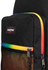 Zaino nero con tasca frontale in rete. Presenta zip con gradiente arcobaleno, hardware metallico e un patch del logo cucito. Tessuto resistente.