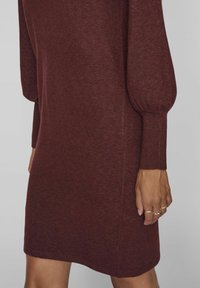 Burgundy svetr šaty s dlouhými, vyboulenými rukávy, žebrovanými manžetami a měkkou texturou, na pozadí v neutrálních barvách s rukou s prsteny.