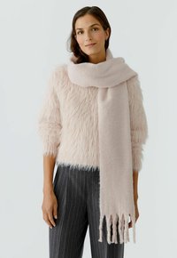 Hellrosa Schal aus weichem, flauschigem Material, um den Hals drapiert, mit fransigen Kanten. Kombiniert mit einem strukturierten, flauschigen Pullover und gestreiften Hosen.