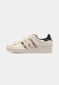 White leather sneaker with a textured, off-white rubber toe cap. Features three brown stripes and a black heel tab. Flat laces and logo detail.