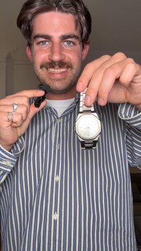 Zilveren polshorloge met een ronde wijzerplaat en metalen band, getoond tegen een meerkleurig gestreept overhemd. Zwarte reversmicrofoon in de linkerhand.