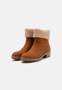 Bottes de cheville en daim marron avec un revers beige en peluche, bout rond et semelle en caoutchouc texturé avec un petit talon empilé.