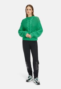 Grüner Strickpullover mit einem Zopfmuster, oversized Schnitt und geripptem Saum, kombiniert mit schwarzen Hosen mit glänzenden Akzenten und sportlichen Sneakers.