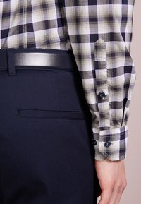 Osoba ubrana w granatowe spodnie, czarny skórzany pasek oraz beżową, białą i czarną w kratkę koszulę z długim rękawem i zapiętymi mankietami.