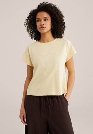 Donna con capelli ricci che indossa una maglietta a maniche corte giallo chiaro e pantaloni larghi marrone scuro, in piedi con una mano in tasca.