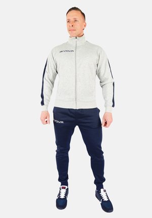 Grijze zip-up sportjack met marineblauwe accenten, gecombineerd met marineblauwe joggers. De stof lijkt glad. Logo zichtbaar op het jack en de broek.