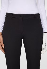 Närbild av svarta tajta byxor med bältesöljer och fickor med dragkedja, bärs tillsammans med en vit långärmad topp och en vit golfhandske på höger hand.