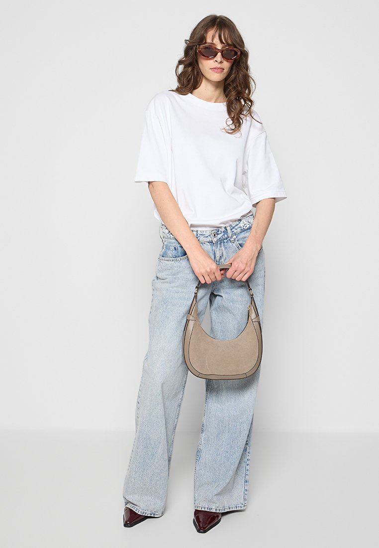 Witte oversized t-shirt, lichtblauwe high-waisted jeans, bruine schoudertas en donkerrode puntige schoenen. Model met golvend haar en zonnebril.
