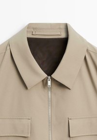 Beige kraagjack met een ritssluiting, voorzien van twee voorzakken en een gladde, gestructureerde stof. Minimalistisch ontwerp met strakke lijnen.