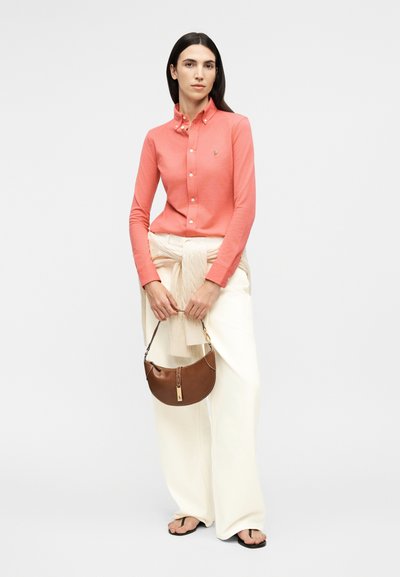 Pikade tumedate juustega naine, kellel on seljas korallivärvi plõksudega särk, vöökohal seotud kampsun, valged laiade säärtega püksid, mustad sandaalid, käes pruun käekott.