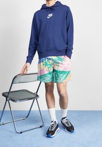 Mörkblå hoodies, mönstrade shorts med palmträd, vita strumpor, svarta sneakers med blå accenter, sittande på en kromstol.