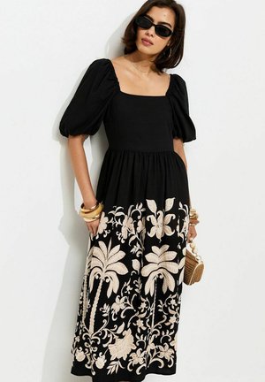 Femme portant une robe noire à manches bouffantes avec broderies beiges de fleurs et de palmiers, lunettes de soleil noires, bracelets dorés, et tenant un petit sac tissé.