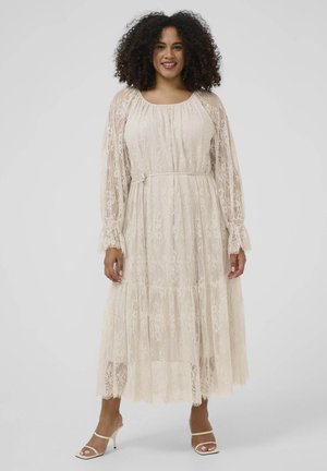 Lächelnde Frau mit lockigem Haar, die ein langes, cremefarbenes Spitzenkleid mit transparenten Ärmeln, einem Taillengürtel und weißen Absatzsandalen trägt.