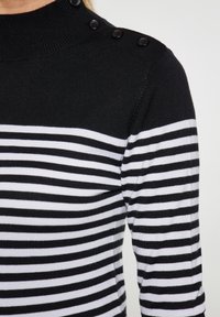 Maglione a righe bianche e nere con collo alto, dotato di bottoni neri sulla spalla e una texture morbida a maglia.