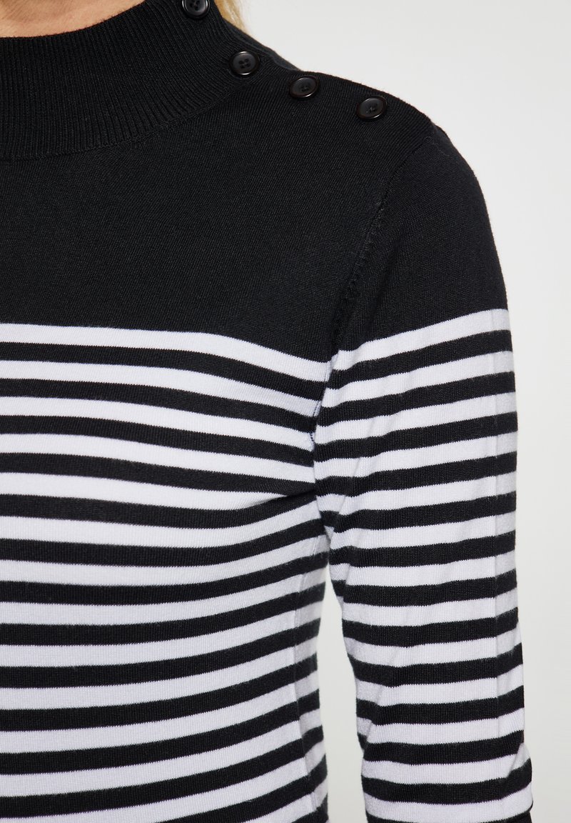 Maglione a righe bianche e nere con collo alto, dotato di bottoni neri sulla spalla e una texture morbida a maglia.