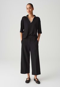 Schwarzes Polo-Hemd mit drei Viertelarmen und weißem Besatz, kombiniert mit weit geschnittenen schwarzen Hose. Das Outfit hat eine lockere Passform und eine weiche Textur.