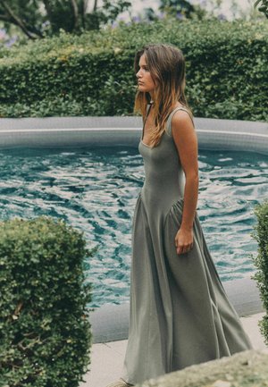 Kvinde i en lang grå kjole, der står ved siden af en cirkulær pool, omgivet af grønne hække og løv i en udendørs havesammenhæng.