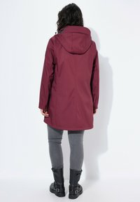 Chaqueta impermeable burdeos con capucha, manga larga y un corte holgado. Presenta una textura suave y bolsillos laterales, combinada con jeans ajustados grises y botas negras.