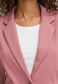 Růžový blazer s texturovanou tkaninou a klopovými límci, kombinovaný s bílým žebrovaným topem. Detailní záběr, který zvýrazňuje výstřih a detaily knoflíků.