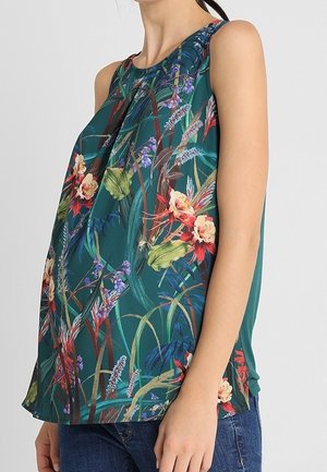 Zielona bluzka bez rękawów z kolorowym kwiatowym i liściastym wzorem, noszona z niebieskimi dżinsami, pokazana z boku na kobiecie.