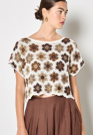 Frau trägt ein braunes und beiges gehäkeltes, sechseckiges Muster-Bauchfreies Oberteil und einen hoch taillierten, braunen, plissierten Rock, schaut nach rechts.