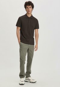 Brown cotton polo shirt with a collar, three buttons, paired with olive green pants and white sneakers with gray accents.