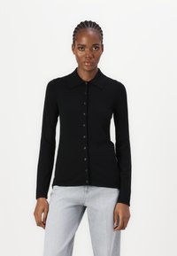 Polo a maniche lunghe nera in maglia con colletto, caratterizzata da una chiusura con bottoni. Il materiale ha una texture liscia, abbinato a jeans grigi chiari.