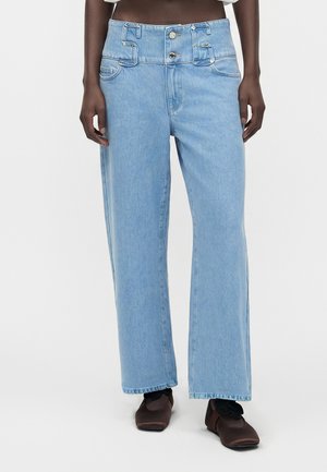Person som har på seg lyseblå jeans med rette ben, dobbelte knapper i livet og brune sko med firkantet tupp mot en ensfarget bakgrunn.