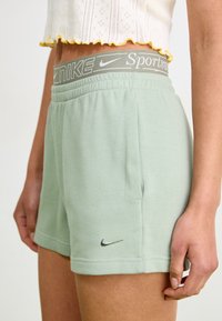 Světle zelené Nike šortky s šedým elastickým pasem, na kterém je text "Nike Sportswear". Měkká látka s stříbrným logem Nike na lemu.