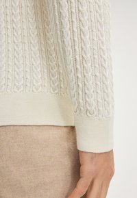 Kabelbrei crème trui met een textuurdesign, geribbelde manchetten en een zachte afwerking. De trui is gelaagd over een beige stof.