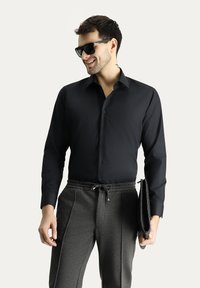 Camicia nera con bottoni e maniche lunghe, abbinata a pantaloni della tuta grigi con coulisse. Modella che tiene una pochette nera e indossa occhiali da sole.