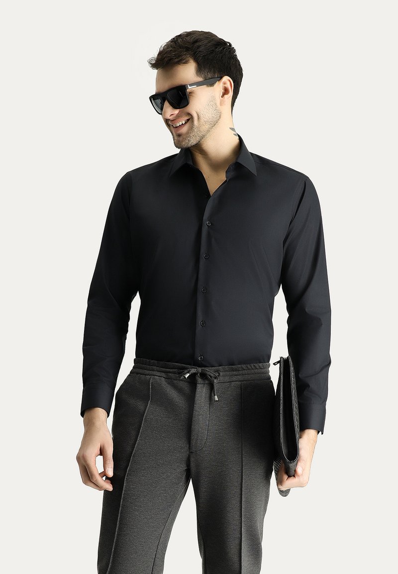 Camicia nera con bottoni e maniche lunghe, abbinata a pantaloni della tuta grigi con coulisse. Modella che tiene una pochette nera e indossa occhiali da sole.