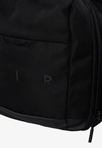 Mochila de tela negra con superficie texturizada, marcas discretas con las letras 'R' e 'I', y un tirador de cremallera negro liso.