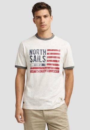 Giovane uomo che indossa una T-shirt bianca e grigia con grafica North Sails rossa e blu e pantaloni beige, in piedi davanti a uno sfondo semplice.