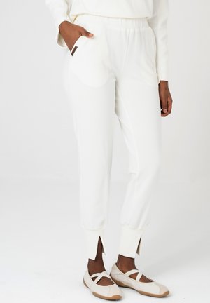 Osoba ubrana w białe spodnie dresowe z bocznymi kieszeniami oraz beżowe płaskie buty z krzyżującymi się paskami, stojąca na jednolitym tle.